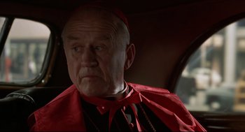 Movie still from “True Confessions” (1981), directed by Ulu Grosbard – An older man wearing a red robe and a red hat; Close Up shot, Over the shoulder angle
