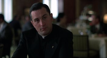 Movie still from “True Confessions” (1981), directed by Ulu Grosbard – A man in a priest's outfit sitting at a table in a restaurant; Close Up shot, Low angle