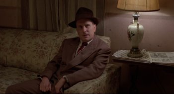 Movie still from “True Confessions” (1981), directed by Ulu Grosbard – A man sitting on a couch wearing a suit and a hat; Medium shot, Low angle