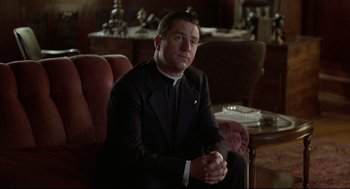 Movie still from “True Confessions” (1981), directed by Ulu Grosbard – A man sitting on a couch wearing a suit and tie; Medium shot, Low angle