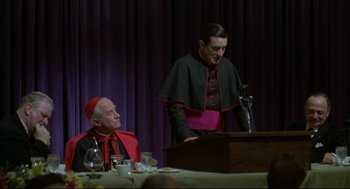 Movie still from “True Confessions” (1981), directed by Ulu Grosbard – A man in a black and red outfit stands at a podium; Medium shot, High angle