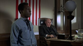 Movie still from “True Grit” (1969), directed by Henry Hathaway – A man sitting at a desk in front of another man in a chair; Medium shot, Low angle