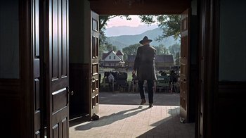Movie still from “True Grit” (1969), directed by Henry Hathaway – A man in a hat and a long coat walking through a doorway; Wide shot, Over the shoulder angle