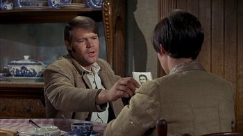 Movie still from “True Grit” (1969), directed by Henry Hathaway – A man holding a picture of a man in front of another man in front of a table; Medium shot, Over the shoulder angle