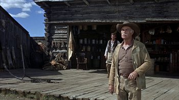 Movie still from “True Grit” (1969), directed by Henry Hathaway – A man in a cowboy hat standing in front of a log cabin; Wide shot, Low angle