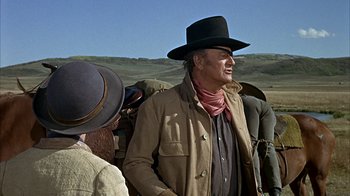 Movie still from “True Grit” (1969), directed by Henry Hathaway – A man wearing a cowboy hat standing in front of a group of people; Medium shot, Over the shoulder angle