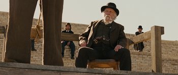 Movie still from “True Grit” (2010), directed by Ethan Coen – An old man sitting on a wooden step with another man sitting on a bench in the background; Wide shot, Low angle