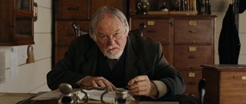 Movie still from “True Grit” (2010), directed by Ethan Coen – An older man sitting at a table writing on a piece of paper; Close Up shot, Over the shoulder angle