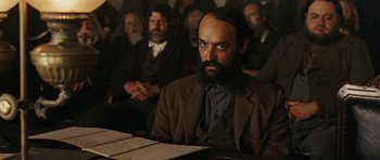 Movie still from “True Grit” (2010), directed by Ethan Coen – A man with a beard sitting in front of a group of people; Medium shot, Low angle