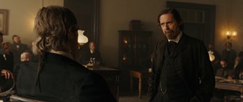 Movie still from “True Grit” (2010), directed by Ethan Coen – A man in a suit and a tie standing next to another man in a suit; Medium shot, Over the shoulder angle