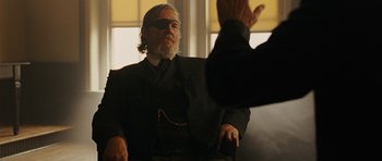 Movie still from “True Grit” (2010), directed by Ethan Coen – An older man wearing a black suit and a blindfold is sitting in a chair; Medium shot, Over the shoulder angle