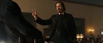 Movie still from “True Grit” (2010), directed by Ethan Coen – A man in a suit and tie holding up a peace sign; Medium shot, Over the shoulder angle