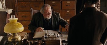 Movie still from “True Grit” (2010), directed by Ethan Coen – An older man sitting at a table with a book; Medium shot, Over the shoulder angle