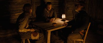 Movie still from “True Grit” (2010), directed by Ethan Coen – A man sitting at a wooden table in a dark room; Medium shot, Over the shoulder angle