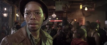 Movie still from “Tsotsi” (2005), directed by Gavin Hood – A man with a hat and glasses in front of a group of people; Close Up shot, Low angle