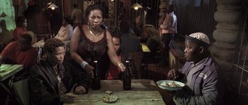 Movie still from “Tsotsi” (2005), directed by Gavin Hood – A woman sitting at a table with two bottles of beer; Medium shot, Over the shoulder angle
