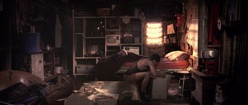 Movie still from “Tsotsi” (2005), directed by Gavin Hood – A man sitting on top of a bed next to a pile of papers; Wide shot, High angle