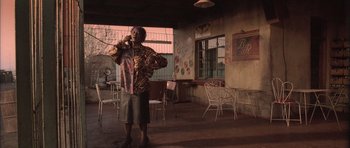 Movie still from “Tsotsi” (2005), directed by Gavin Hood – A man standing in front of a building with a cell phone to his ear; Wide shot, Low angle