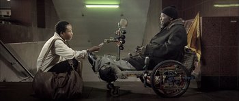 Movie still from “Tsotsi” (2005), directed by Gavin Hood – A man sitting in a wheel chair in a room with a woman; Wide shot, Low angle