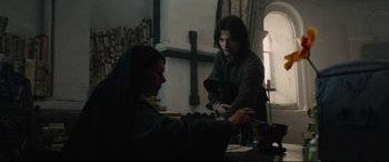 Movie still from “Tulip Fever” (2017), directed by Justin Chadwick – Two people sitting at a table in front of a cross on the wall; Medium shot, Over the shoulder angle