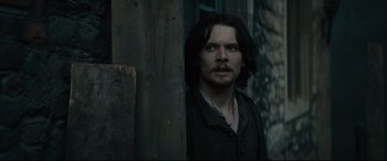 Movie still from “Tulip Fever” (2017), directed by Justin Chadwick – A man with long black hair standing next to a wooden wall; Medium shot, Low angle