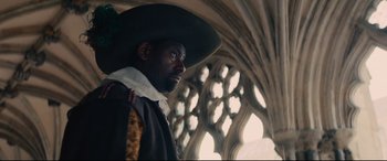 Movie still from “Tulip Fever” (2017), directed by Justin Chadwick – A man wearing a hat standing in front of a window; Medium shot, Low angle