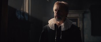 Movie still from “Tulip Fever” (2017), directed by Justin Chadwick – A man with a beard wearing a black and white collar; Close Up shot, Over the shoulder angle