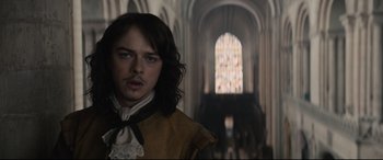 Movie still from “Tulip Fever” (2017), directed by Justin Chadwick – A man with long brown hair standing in front of a stained glass window; Close Up shot, Over the shoulder angle