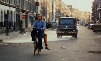 Movie still from “Turkish Delight” (1973), directed by Paul Verhoeven – A woman riding a bike down a street with a van behind it; Wide shot, Low angle