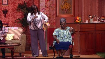 Movie still from “Madea on the Run” (2017), directed by Derrick Doose – Two women in a living room one is holding a banana and the other is sitting on a chair; Wide shot, High angle