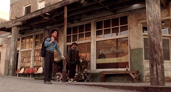 Movie still from “U Turn” (1997), directed by Oliver Stone – A man standing next to another man in front of a building; Wide shot, Low angle