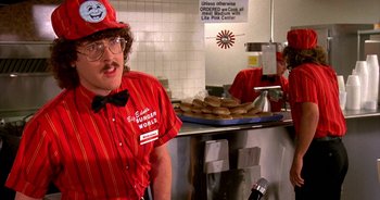 Movie still from “UHF” (1989), directed by Jay Levey – A man in a red and white uniform standing in front of donuts; Medium shot, Over the shoulder angle