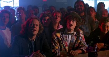 Movie still from “UHF” (1989), directed by Jay Levey – A group of people sitting in front of a crowd; Medium shot, Over the shoulder angle