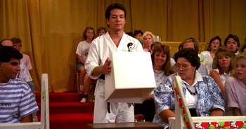 Movie still from “UHF” (1989), directed by Jay Levey – A man holding a box in front of a group of people; Medium shot, Over the shoulder angle