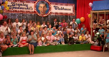 Movie still from “UHF” (1989), directed by Jay Levey – A group of people sitting on a stage in front of a sign; Wide shot, High angle