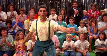Movie still from “UHF” (1989), directed by Jay Levey – A group of people sitting and standing in front of a man; Medium shot, Low angle