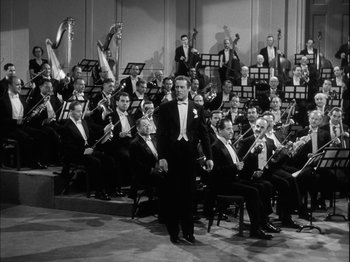 Movie still from “Unfaithfully Yours” (1948), directed by Preston Sturges – A man in a black suit standing in front of a group of people; Wide shot, High angle