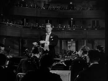 Movie still from “Unfaithfully Yours” (1948), directed by Preston Sturges – A man in a tuxedo is conducting an orchestra in front of an audience; Wide shot, High angle