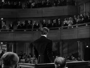 Movie still from “Unfaithfully Yours” (1948), directed by Preston Sturges – A man standing in front of an audience in front of an orchestra; Wide shot, Over the shoulder angle