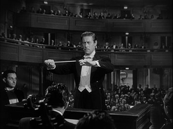 Movie still from “Unfaithfully Yours” (1948), directed by Preston Sturges – A man in a black suit and white bow tie holding a conductor's baton in front of an audience; Medium shot, High angle