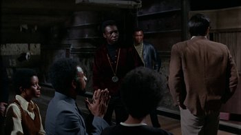 Movie still from “Uptight” (1968), directed by Jules Dassin – A group of people gathered around a man in a red shirt; Medium shot, Over the shoulder angle