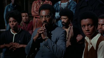 Movie still from “Uptight” (1968), directed by Jules Dassin – A group of people standing around a man in a suit; Medium shot, Over the shoulder angle