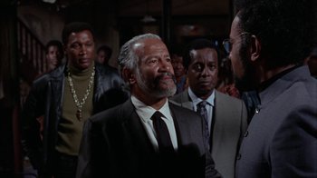 Movie still from “Uptight” (1968), directed by Jules Dassin – An older man wearing a suit and tie in front of a group of men; Medium shot, Over the shoulder angle