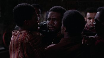 Movie still from “Uptight” (1968), directed by Jules Dassin – A group of men standing next to each other in a dark room; Medium shot, Over the shoulder angle