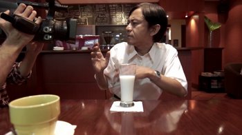 Movie still from “V/H/S/2” (2013), directed by Timo Tjahjanto – A person sitting at a table with a glass of milk; Medium shot, Over the shoulder angle