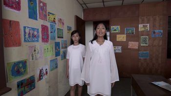 Movie still from “V/H/S/2” (2013), directed by Timo Tjahjanto – Two young girls standing in a room with a wall of children's drawings on the wall behind them; Medium shot, Low angle