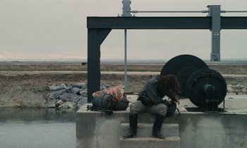 Movie still from “Vagabond” (1985), directed by Agnès Varda – A man sitting on a concrete wall next to a body of water; Wide shot, Low angle