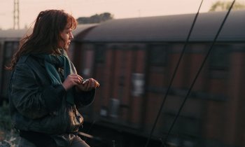 Movie still from “Vagabond” (1985), directed by Agnès Varda – A woman standing on a train platform looking at her cell phone; Medium shot, Over the shoulder angle