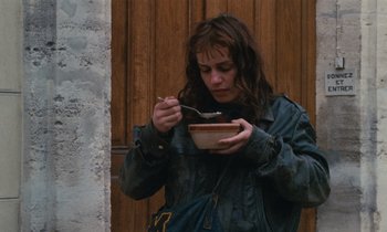 Movie still from “Vagabond” (1985), directed by Agnès Varda – A woman eating a bowl of food while standing in front of a building; Medium shot, High angle