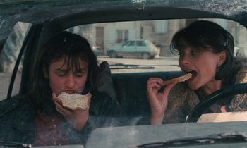 Movie still from “Vagabond” (1985), directed by Agnès Varda – Two women are sitting in the back of a car eating food; Medium shot, Over the shoulder angle